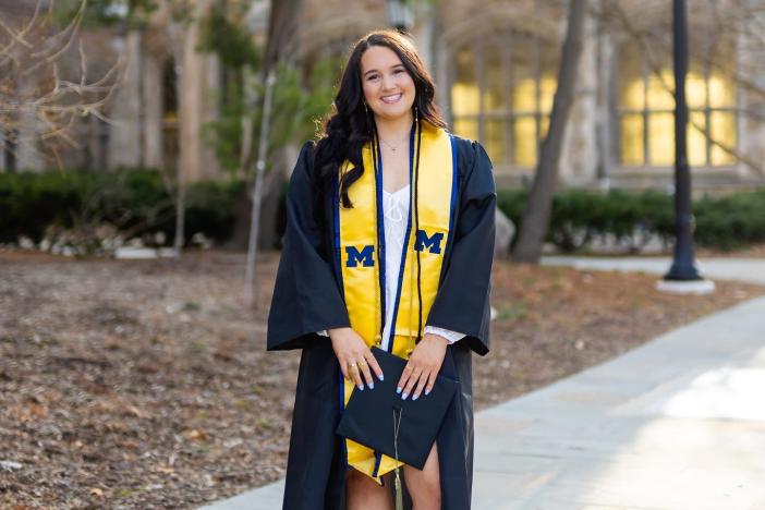 Noelle Bedard stands on the sidewalk in front of central campus in her graduation robe and a yellow stole with blue block Ms, holding her graduation cap.