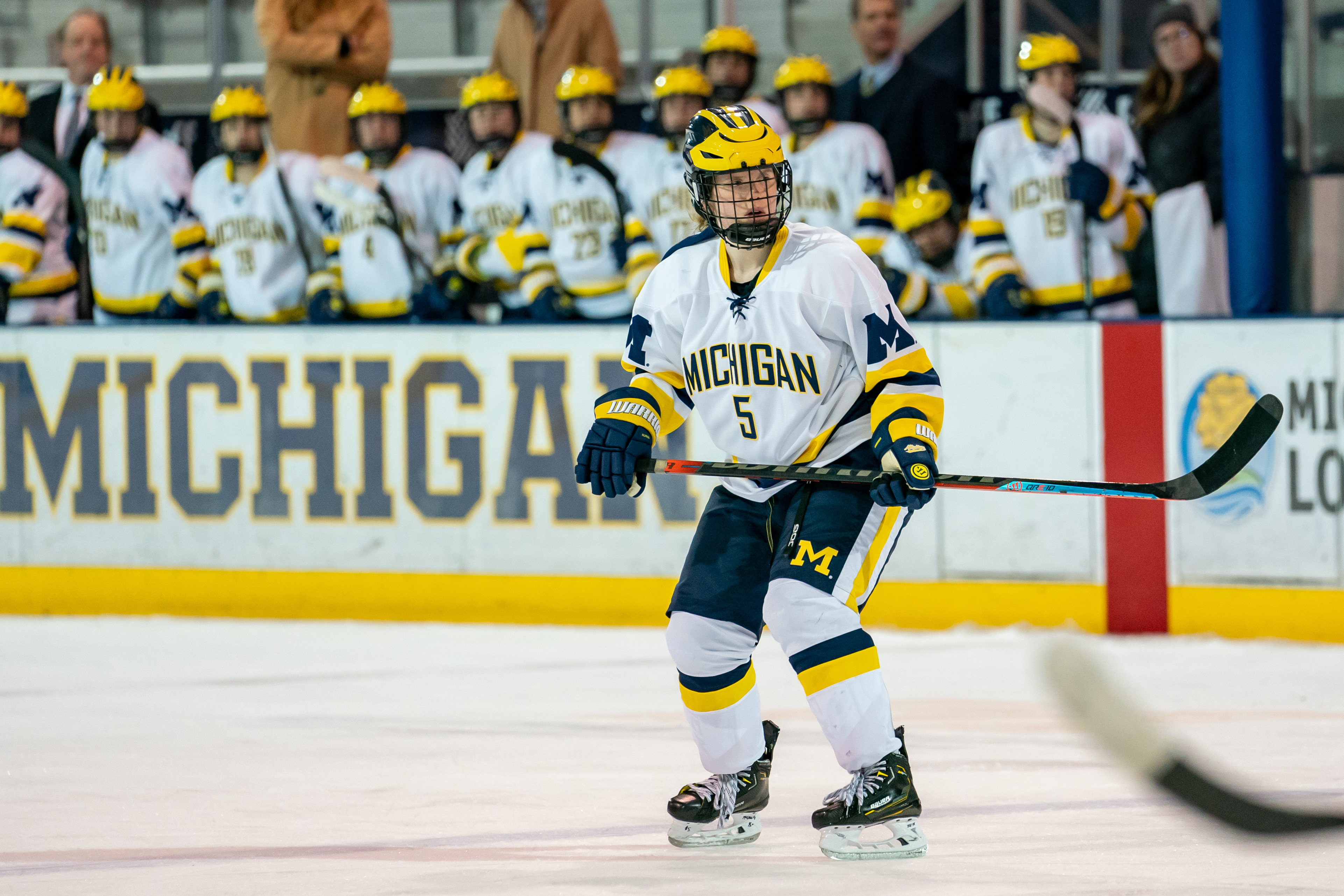 A woman in a white, blue, and yellow Michigan hockey uniform, holding a stick on the ice.
