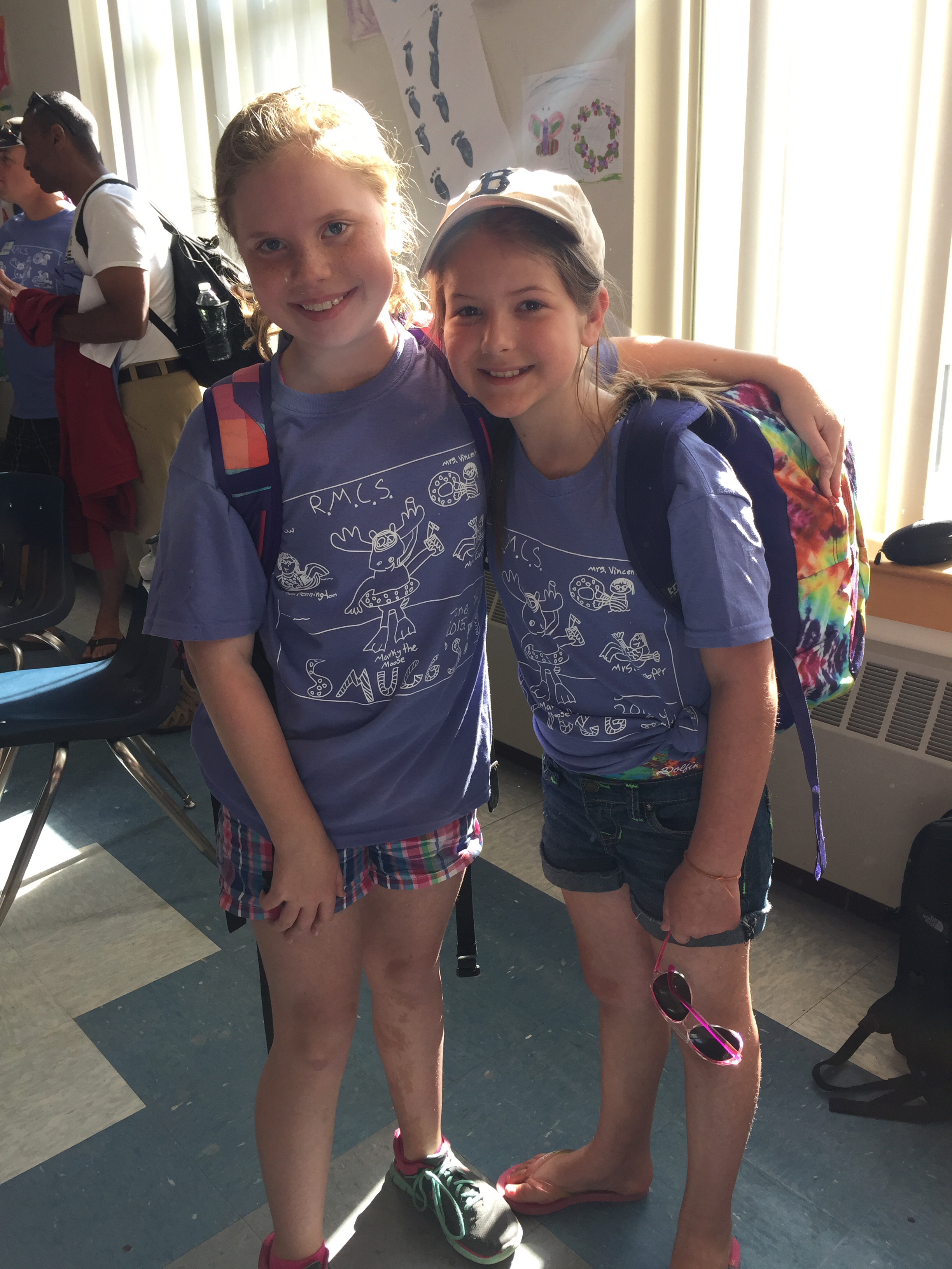 Two young girls in purple T-shirts with a white design, wearing backpacks.