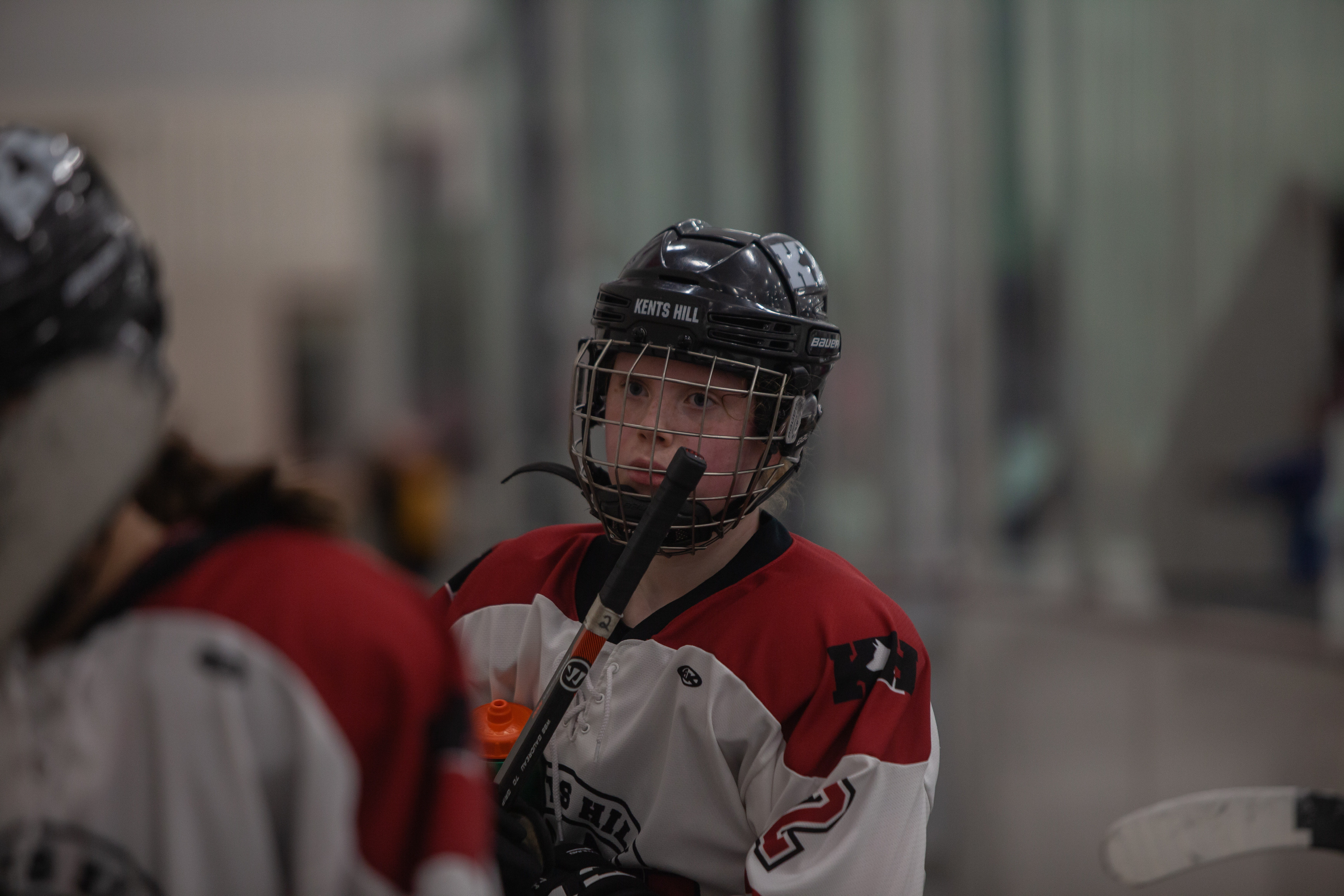 A teenage woman in a red and white hockey uniform with a black helmet and a black stick.