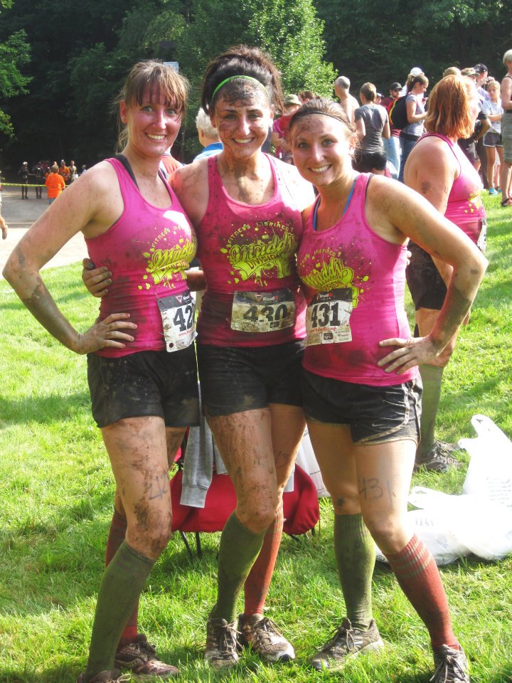 Three people in pink tank tops, black athletic shorts, knee high socks, and dark tennis shoes, all splattered in mud, pose at the end of a race.