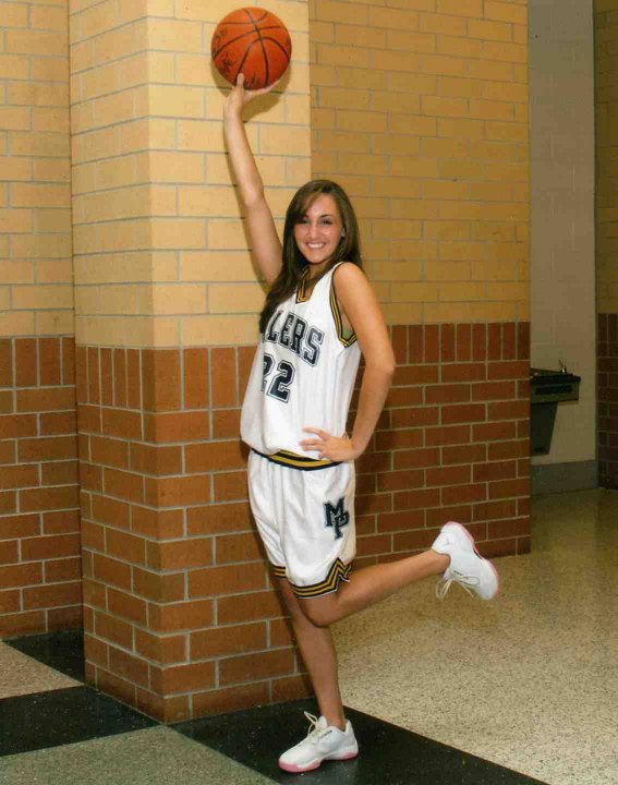 A person in a white basketball uniform holding a basketball above their head. The person has long straight brown hair.