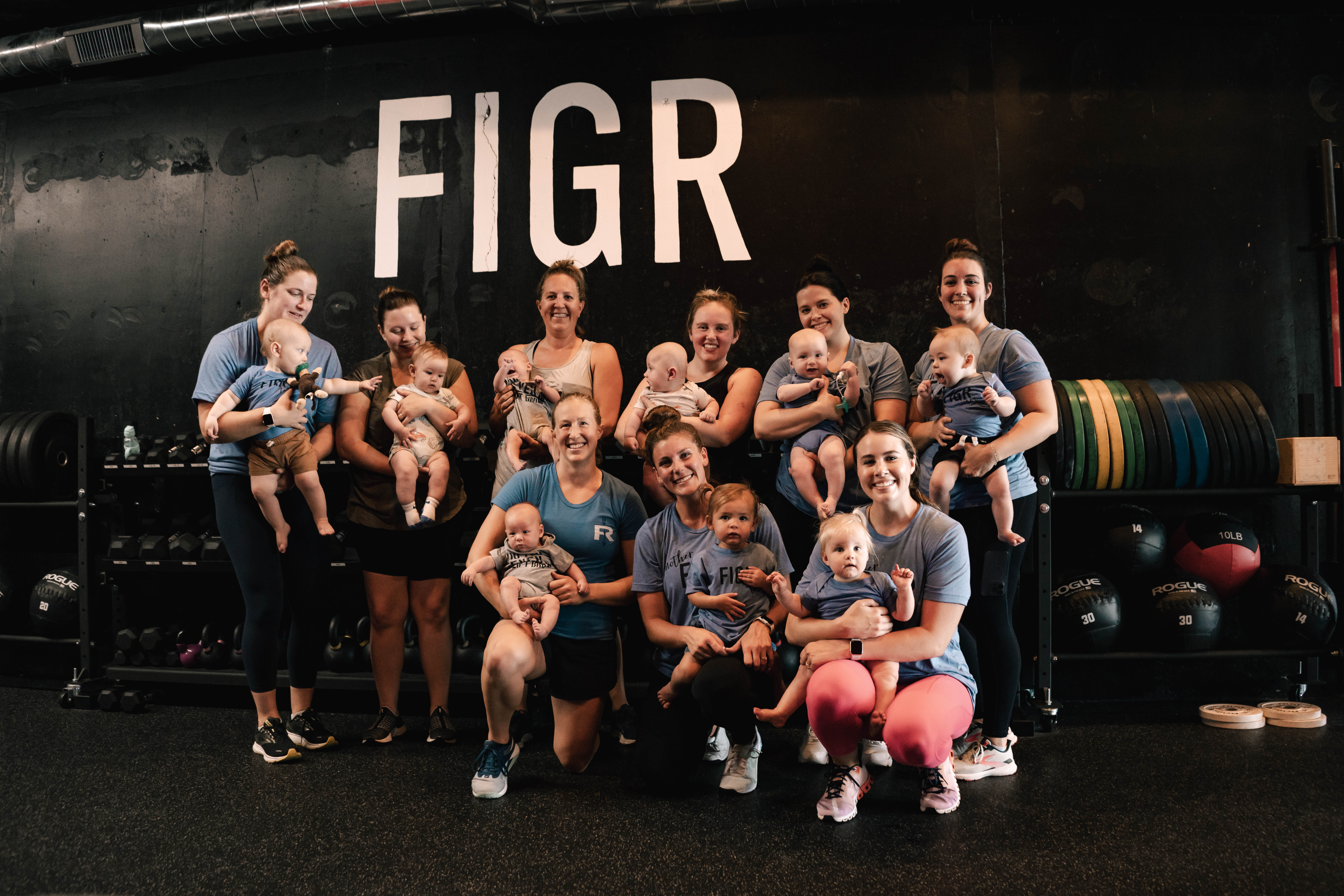 A group of moms in workout clothing holding their babies, all standing in front of a black wall with the word "FIGR" in tall white lettering.