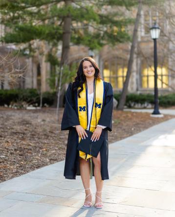 Noelle Bedard stands on the sidewalk in front of central campus in her graduation robe and a yellow stole with blue block Ms, holding her graduation cap.