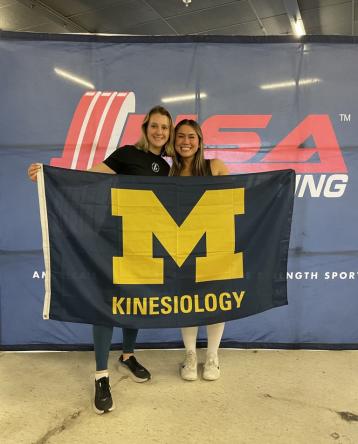Julianna King stands with one of the powerlifters she coaches at a meet, holding a Kinesiology flag
