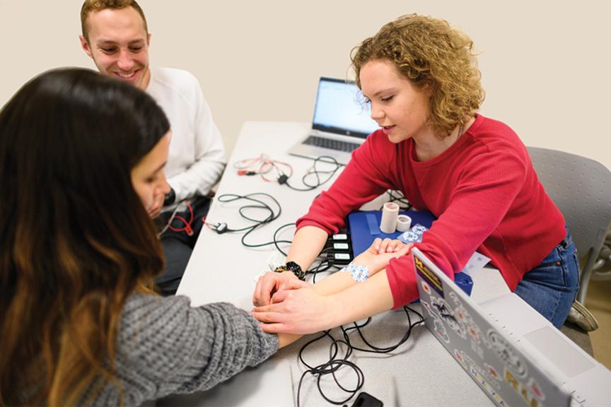 Three students sitting around a table connecting sensors to a student&#039;s arm.