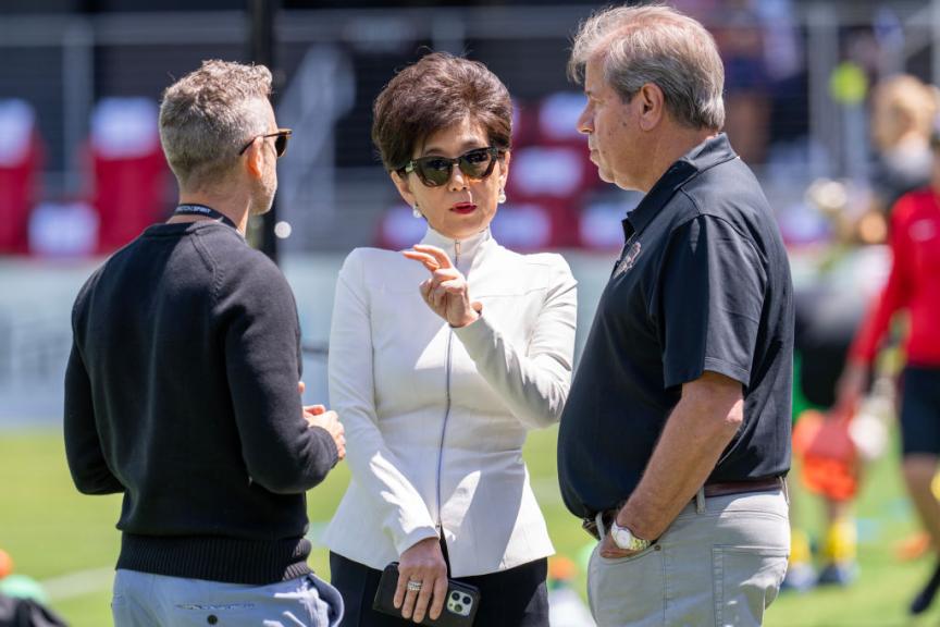 An Asian woman with short brown hair wearing a white jacket with a silver zipper up the front, sunglasses, pearl earrings, and bright pink lipstick stands on a soccer field and speaks with two men in black shirts.