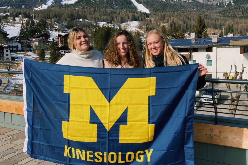 Hannah Janovsky, Hallie Zlotnik, and Jordin Rickard, all sport management students, show off a Kinesiology flag in front of the Dolomites in northern Italy