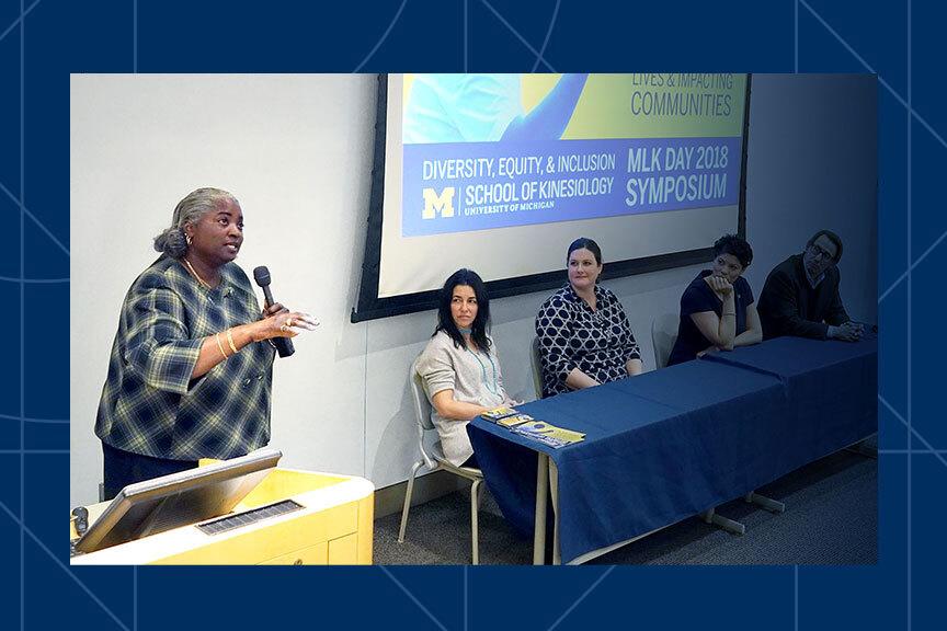 Dr. Ketra Armstrong, a Black woman with grey hair in a plaid jacket, stands at the front of a room holding a microphone. Next to her is a table with four people who are turned toward her. Behind them is a projection screen with the phrases, &quot;Diversity, Equity &amp; Inclusion,&quot; &quot;MLK Day 2018 Symposium,&quot; and &quot;School of Kinesiology.&quot;