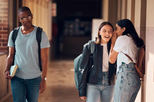 A Black teenage boy walks down a school hallway as two white girls whisper.