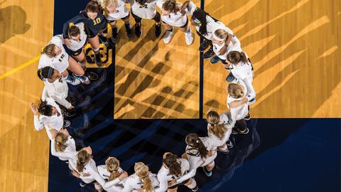 University of Michigan women&#039;s volleyball team linking arms and forming a circle on the U-M volleyball floor.