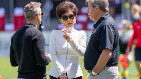 An Asian woman with short brown hair wearing a white jacket with a silver zipper up the front, sunglasses, pearl earrings, and bright pink lipstick stands on a soccer field and speaks with two men in black shirts.