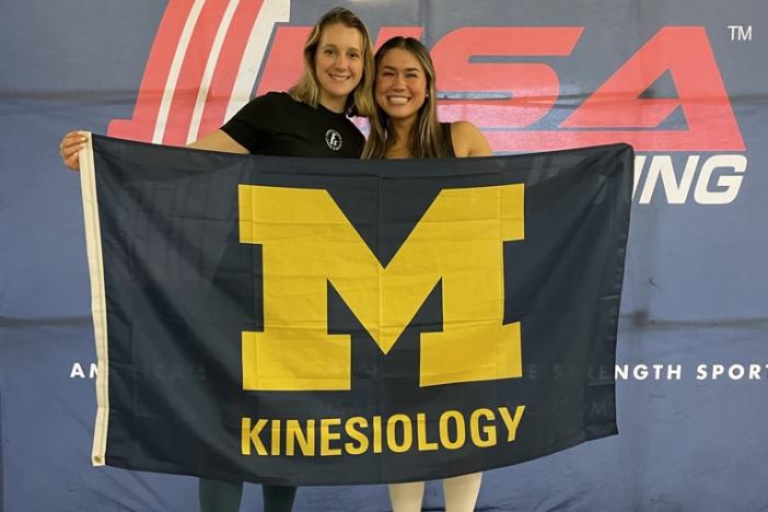 Julianna King stands with one of the powerlifters she coaches at a meet, holding a Kinesiology flag